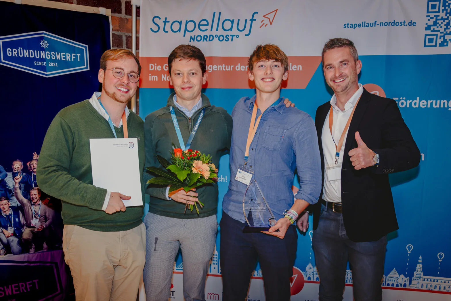 Preisträgerteam Solentil des Businessplanwettbewerb MV 2023 mit Urkunde, Blumenstrauß und Glaspokal vor der Stapellauf- und Gründungswerft-Wand.